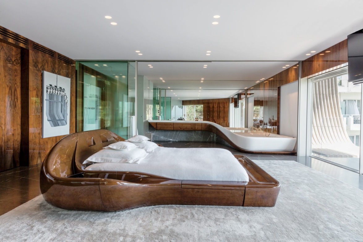 The master bedroom has been furnished with a bed set in a sculpted wood base. The bathroom—with its organically shaped tub and burl walnut-veneer cabinetry—is completely open to the rest of the space. The work of art on the wall is by the Greek artist Yannis Gaitis.