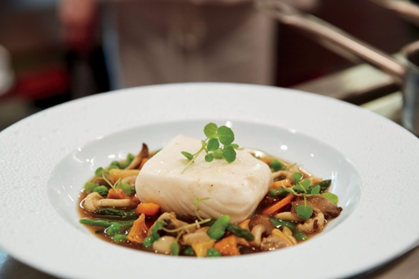 A gourmet dish featuring a piece of white fish atop a bed of mixed vegetables and herbs on a white round plate.