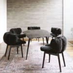 Modern dining room with black cushioned chairs, stone table on a patterned rug, set against a light stone wall and window.