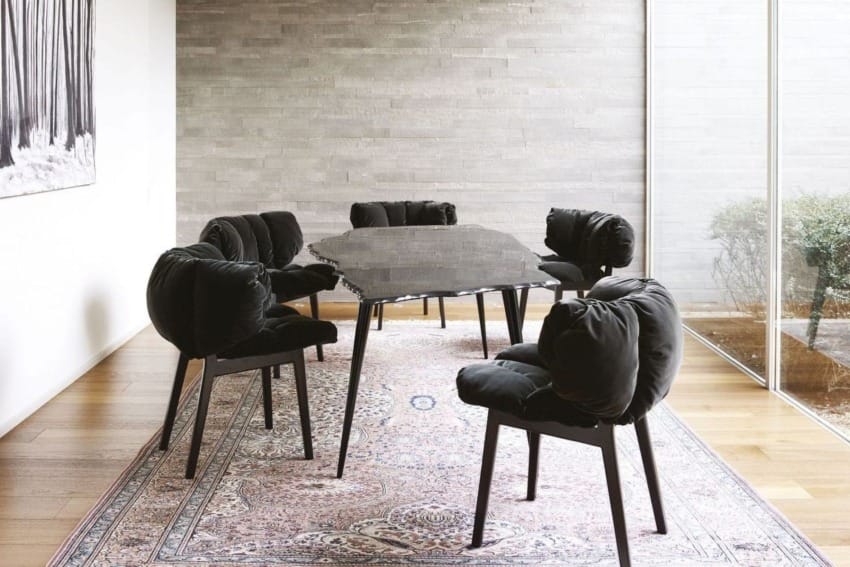 Modern dining room with black cushioned chairs, stone table on a patterned rug, set against a light stone wall and window.