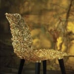 Intricately designed transparent chair with golden hues and black legs against a textured yellow and brown background.