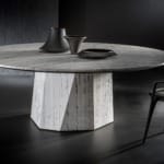 Modern round marble table with geometric base, black chair, and decorative vases on top, set against a dark background.