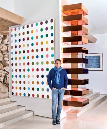 Person standing beside a large colorful dot painting and modern stacked sculpture in a contemporary interior setting.
