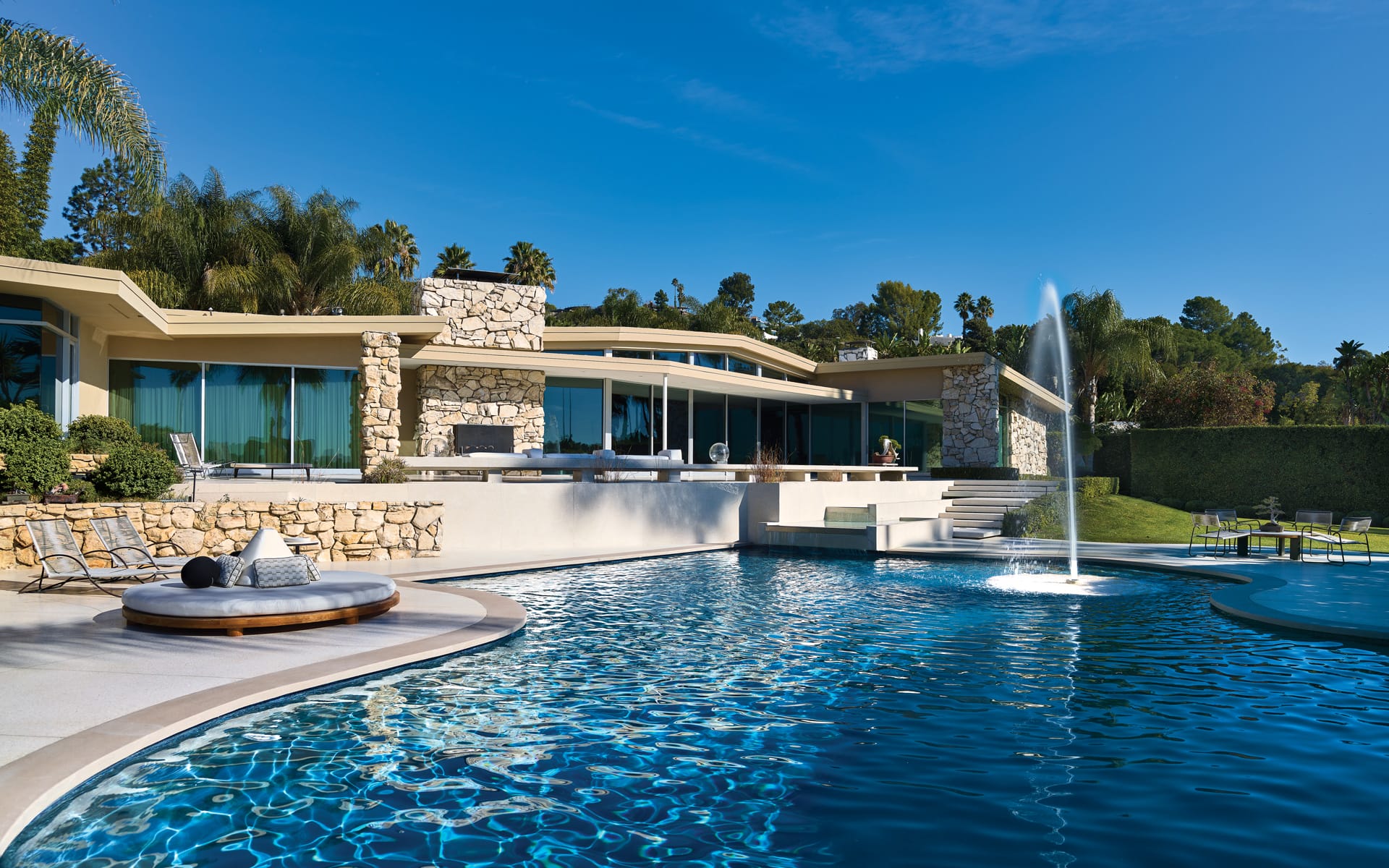 Luxury home with modern architecture, large glass windows, surrounded by lush greenery, and a sparkling swimming pool in front.