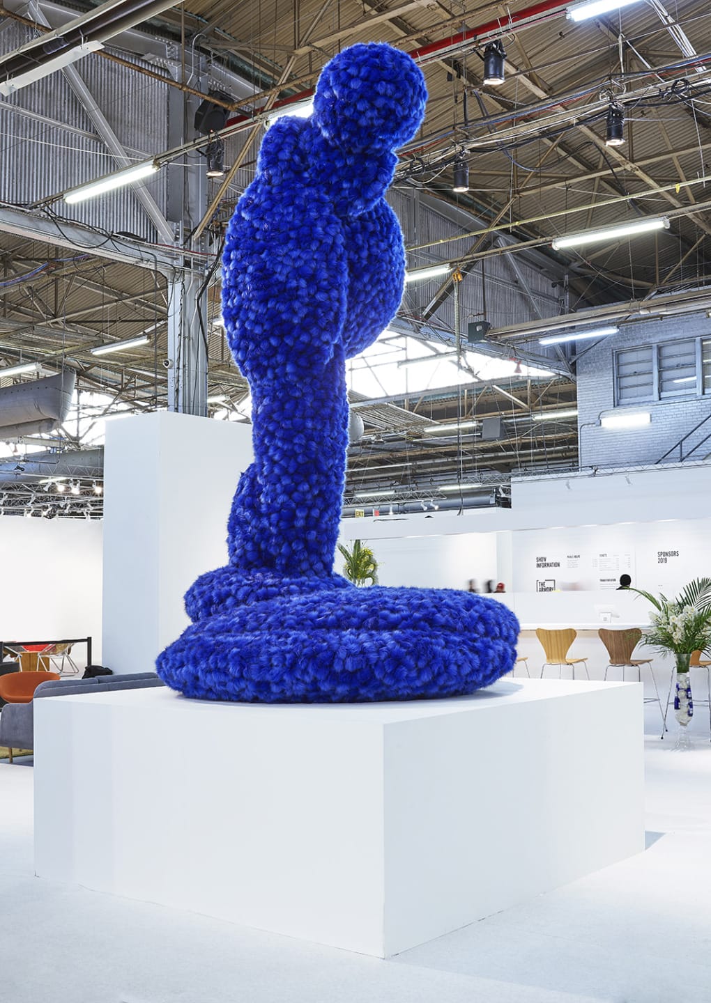 Blue abstract sculpture on a white pedestal in an industrial-style exhibition space with high ceilings and exposed beams.