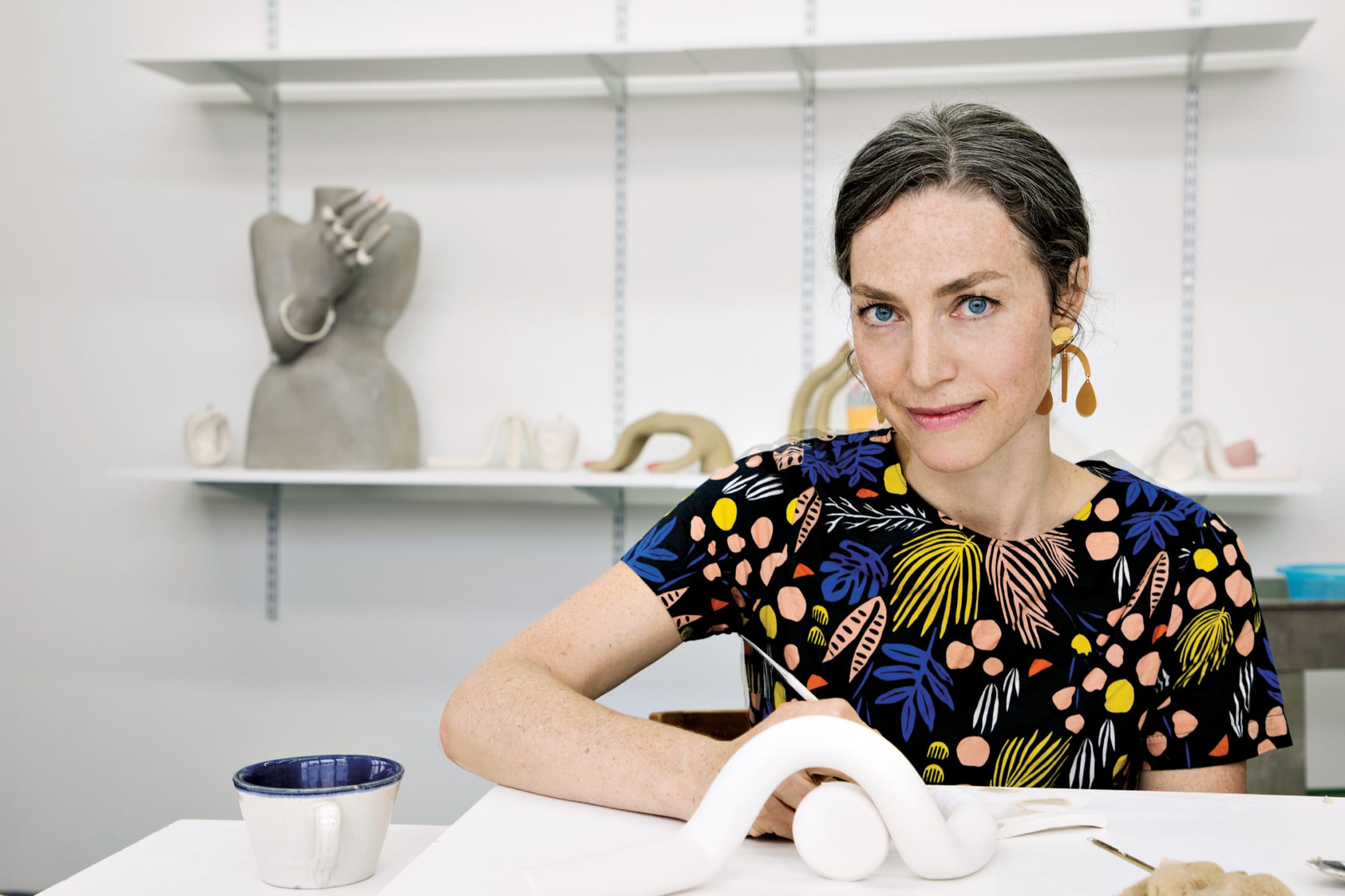 Person with colorful shirt sitting at a table in a studio with ceramics and sculptures displayed on shelves behind.