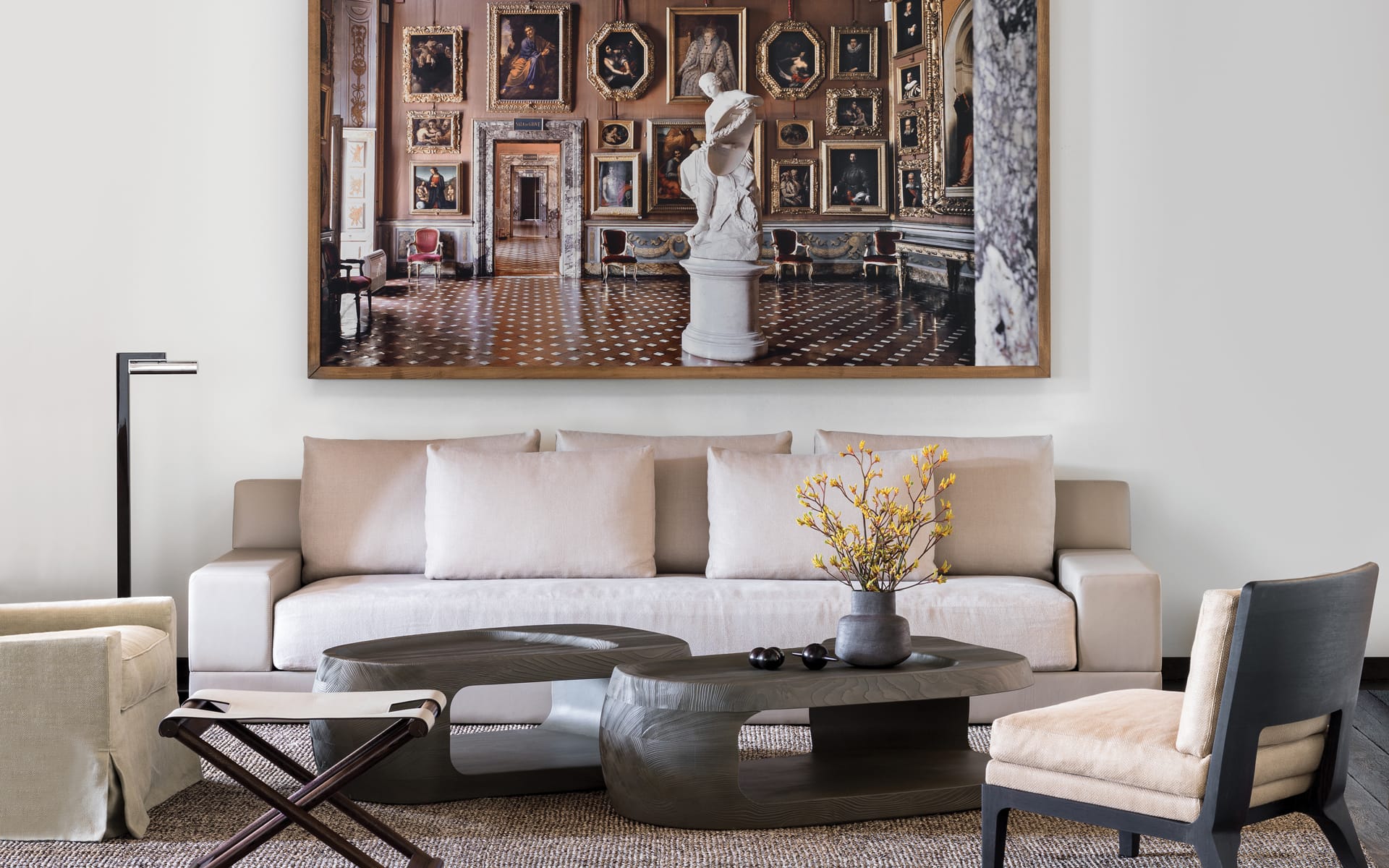 Modern living room with a white sofa, abstract coffee tables, a large painting, and a sculpture in a gallery-inspired setting.