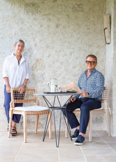 Two people sitting at a small outdoor table with a stone wall background, one reading a newspaper, the other standing nearby.