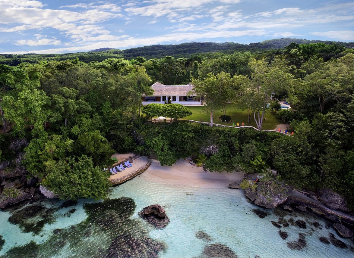 Aerial view of a house surrounded by lush greenery on a coastline with clear blue water and a small sandy beach.