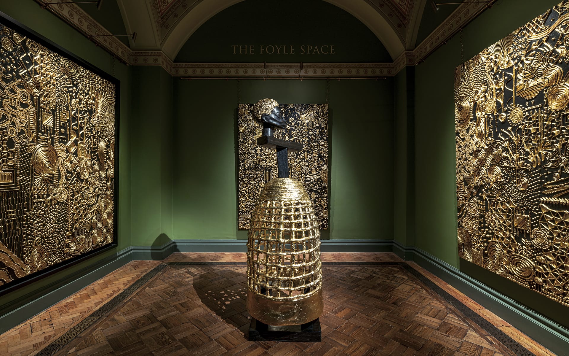 Gold sculptures and intricate panels in an art gallery with green walls and wooden floors.
