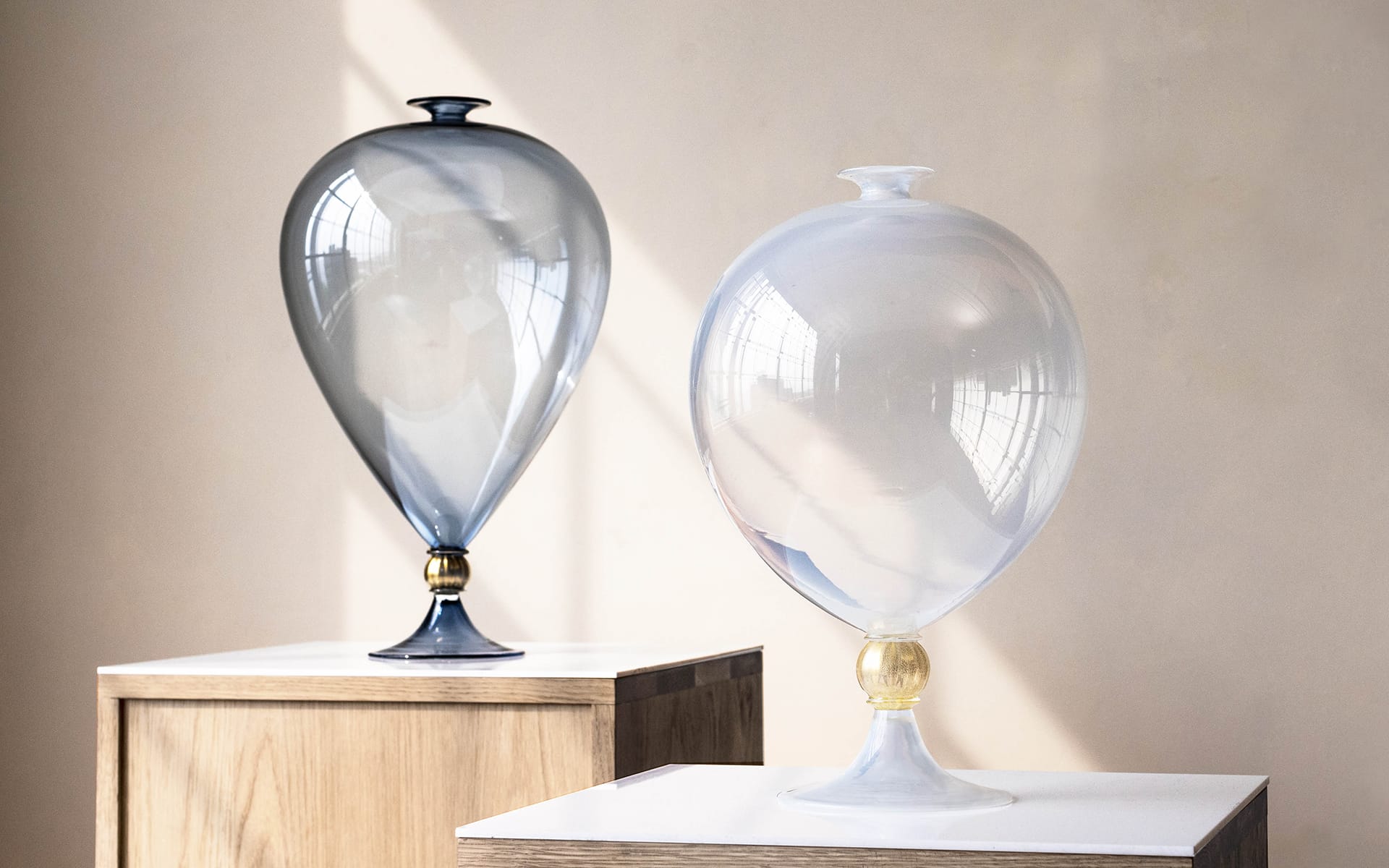 Elegant glass sculptures on wooden stands, featuring smooth, rounded shapes with a reflective surface in an art gallery setting.
