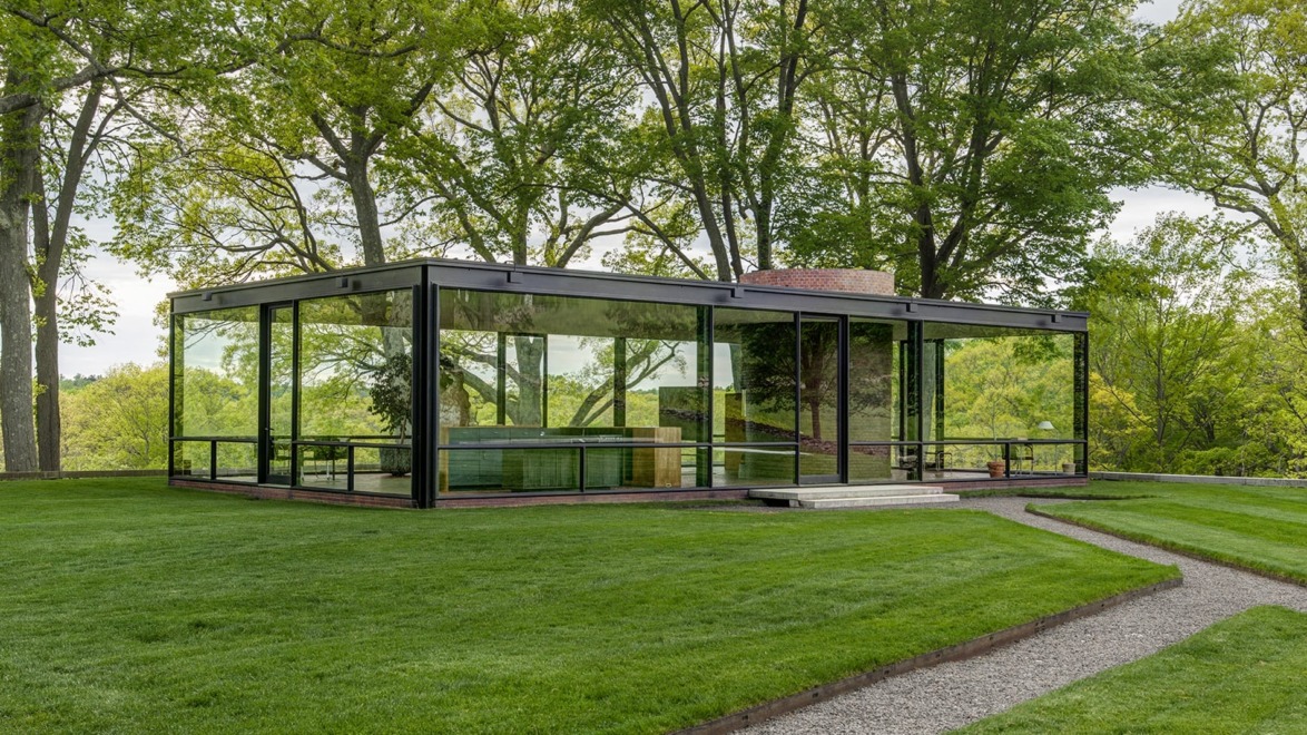 Modern glass house with sleek design, surrounded by lush green trees and well-maintained lawn on a sunny day.