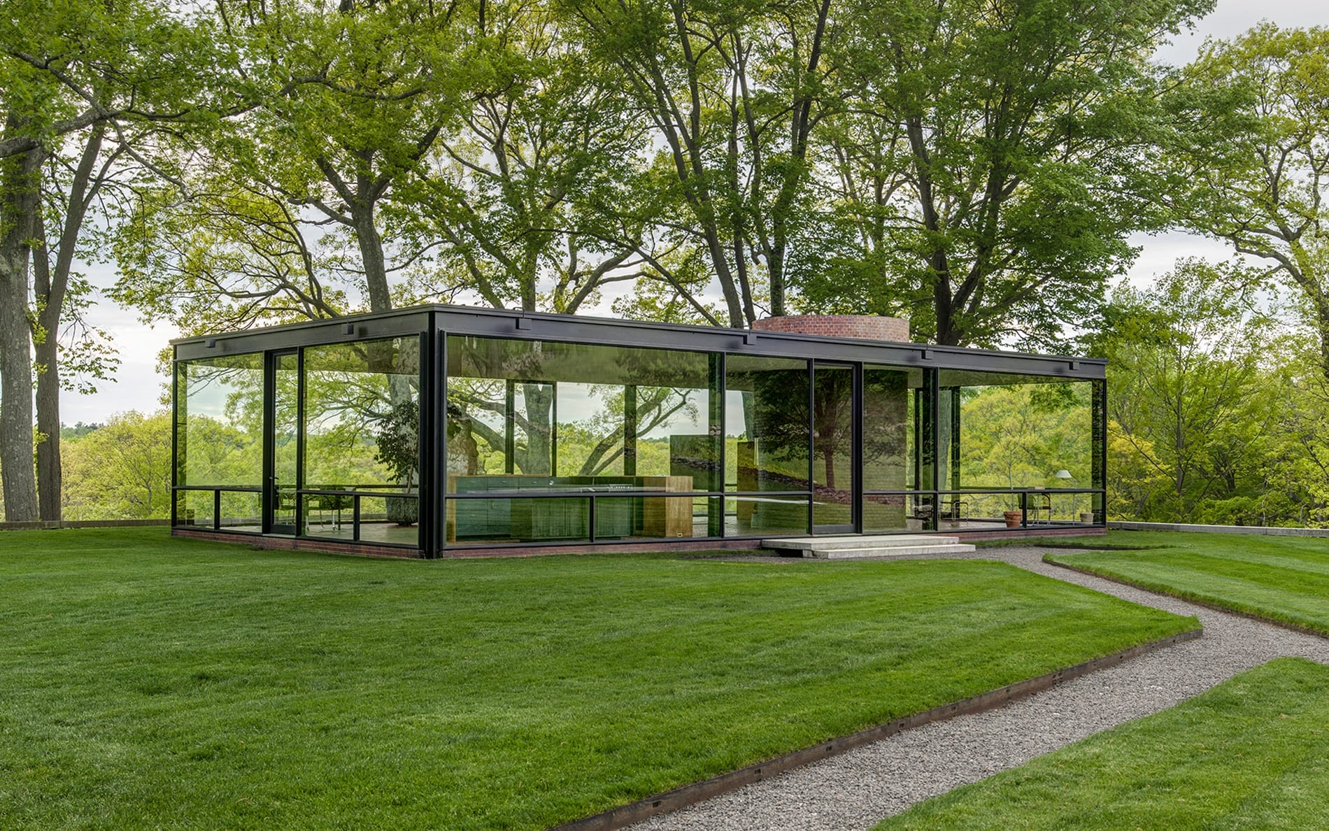 Modern glass house with sleek design, surrounded by lush green trees and well-maintained lawn on a sunny day.