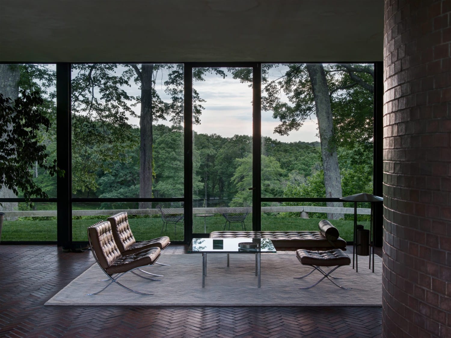 Modern glass-walled living room with leather furniture and forest view, showcasing sleek design and natural surroundings.