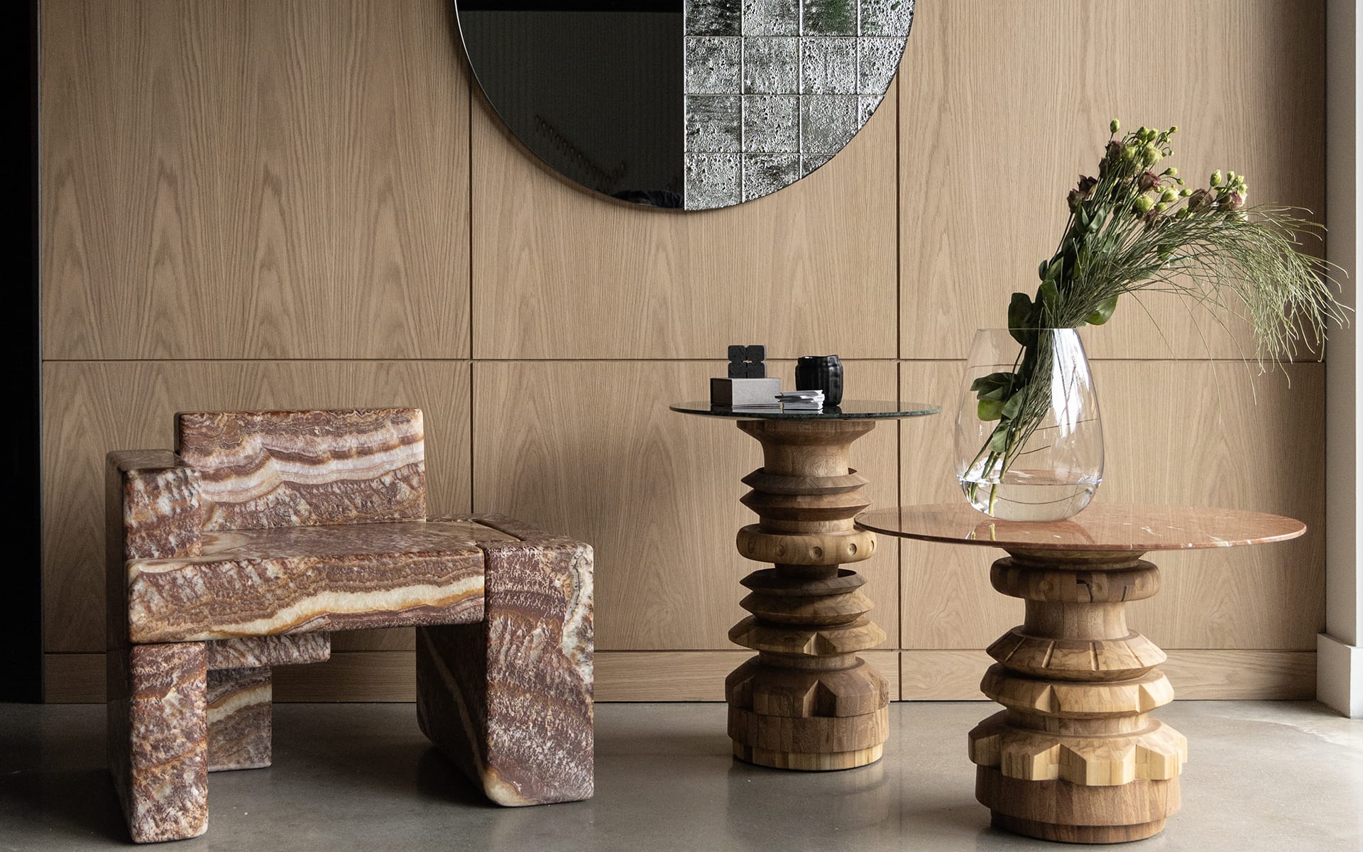 Modern interior with sculptural chair, round mirror, wooden tables, and a vase with flowers in a contemporary setting.