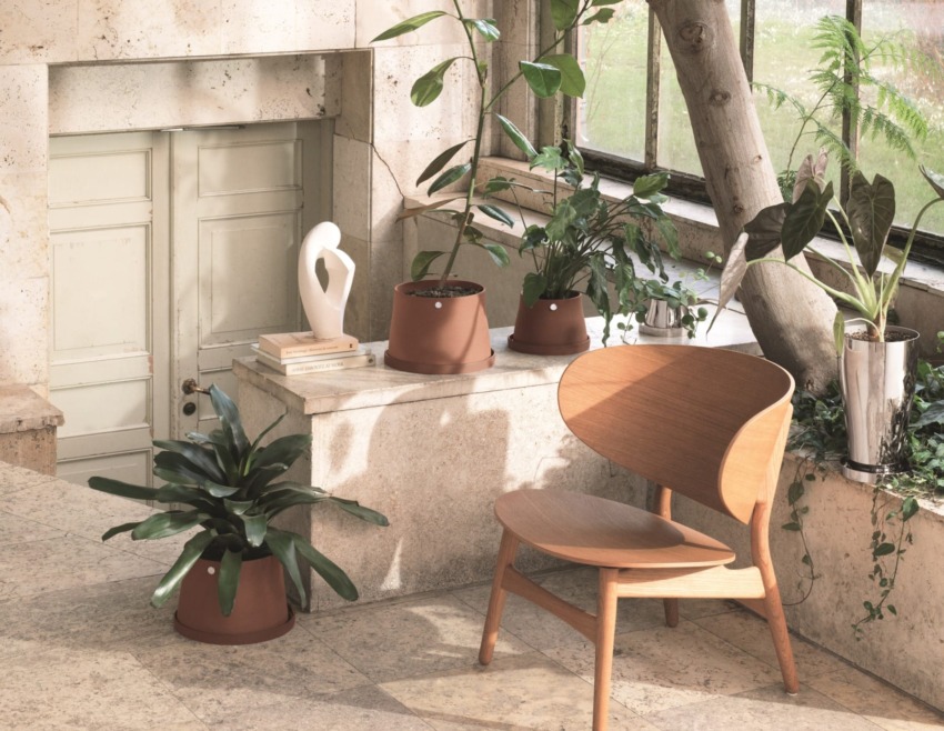 Modern indoor garden scene with wooden chair, potted plants, large windows, and a decorative sculpture on a stone ledge.