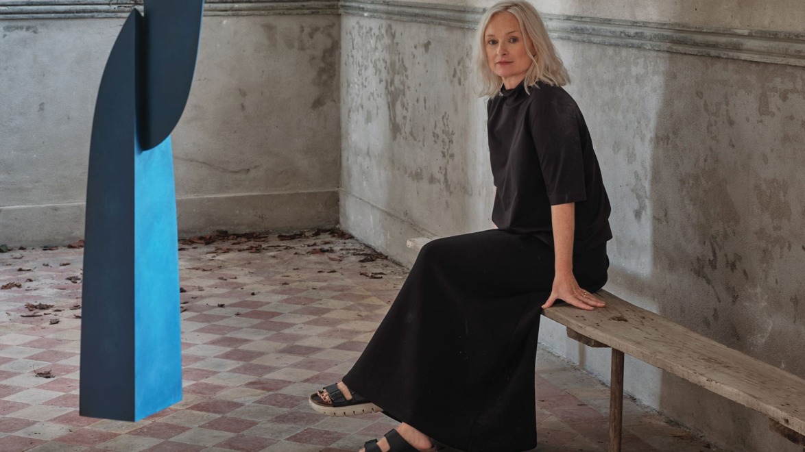 A woman with gray hair sitting on a bench in a room with tiled floor, near a large blue abstract sculpture.