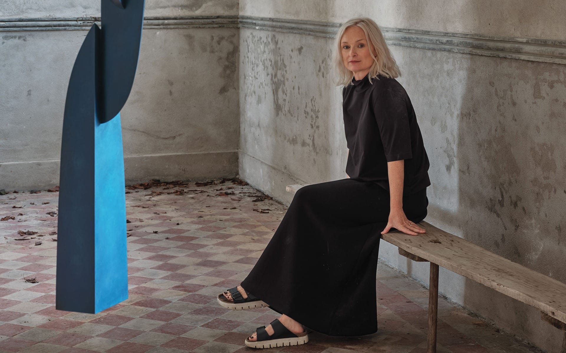 A woman with gray hair sitting on a bench in a room with tiled floor, near a large blue abstract sculpture.