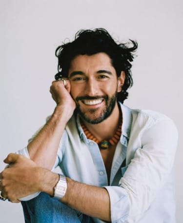 Smiling man with curly hair and a beaded necklace, wearing a light blue shirt, sitting against a plain background.