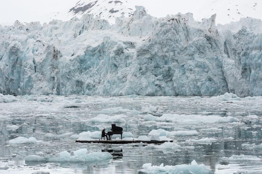 Person playing piano on floating platform surrounded by icebergs and snowy mountains.