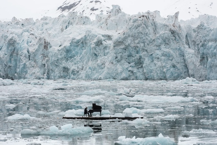Person playing piano on floating platform surrounded by icebergs and snowy mountains.