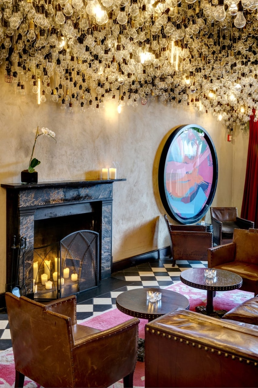 Cozy lounge with antique fireplace, leather chairs, ornate light-filled ceiling, and round window with abstract art.