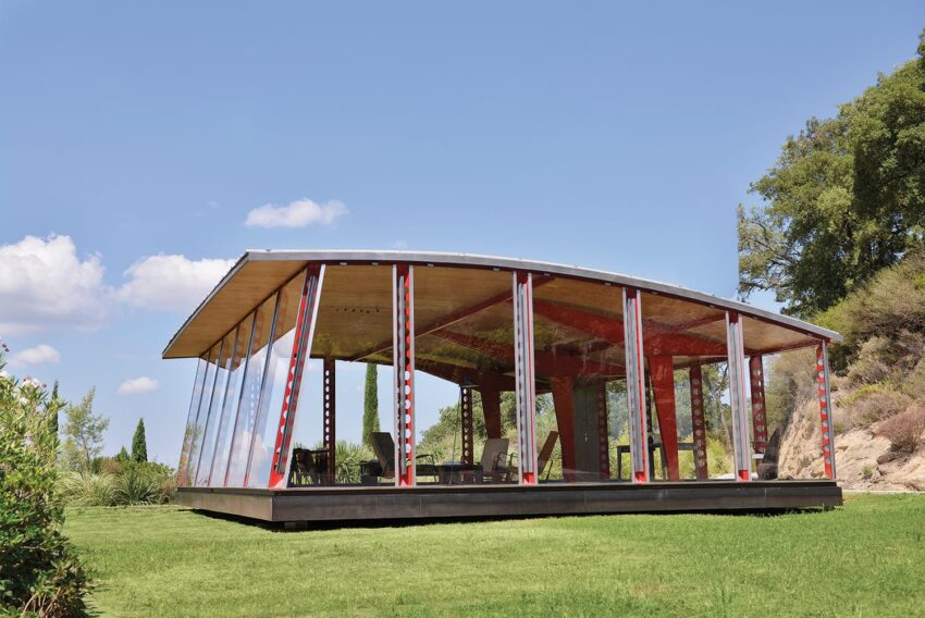 Modern glass-walled pavilion on grass with trees and cloudy sky in the background.