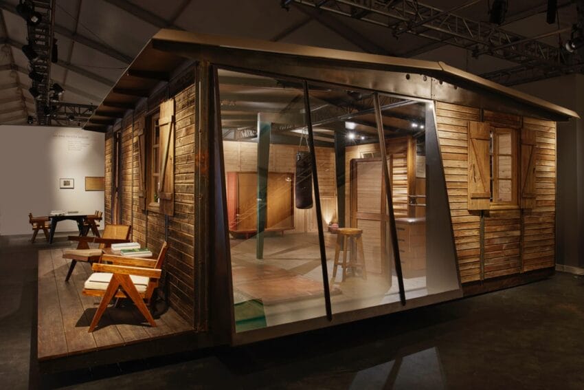 Wooden exhibition structure with modern design, featuring large windows and wooden furniture displayed in an artistic setting.