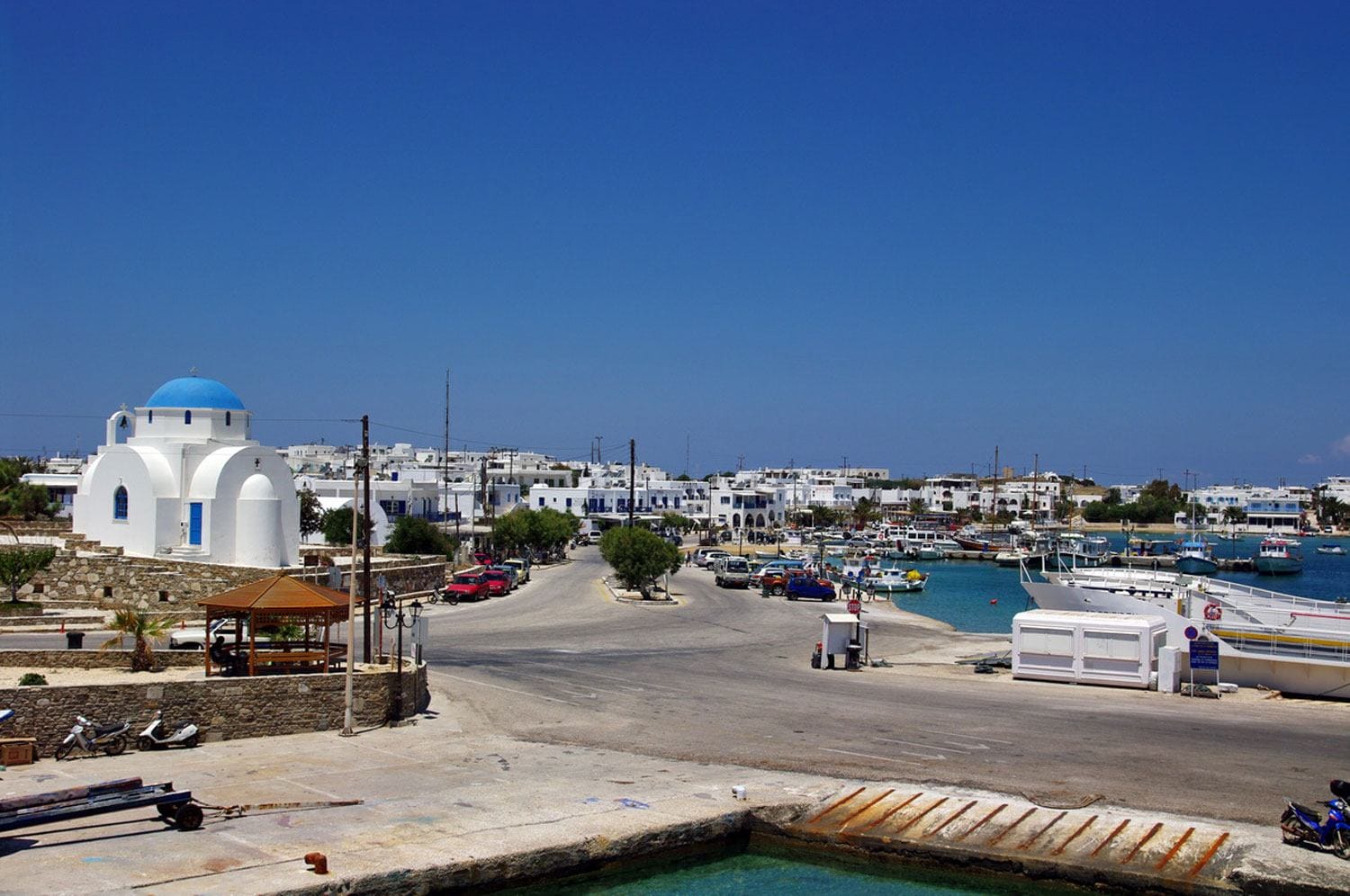 Scenic view of a coastal Greek village with white buildings, a blue-domed church, and a small harbor with boats.