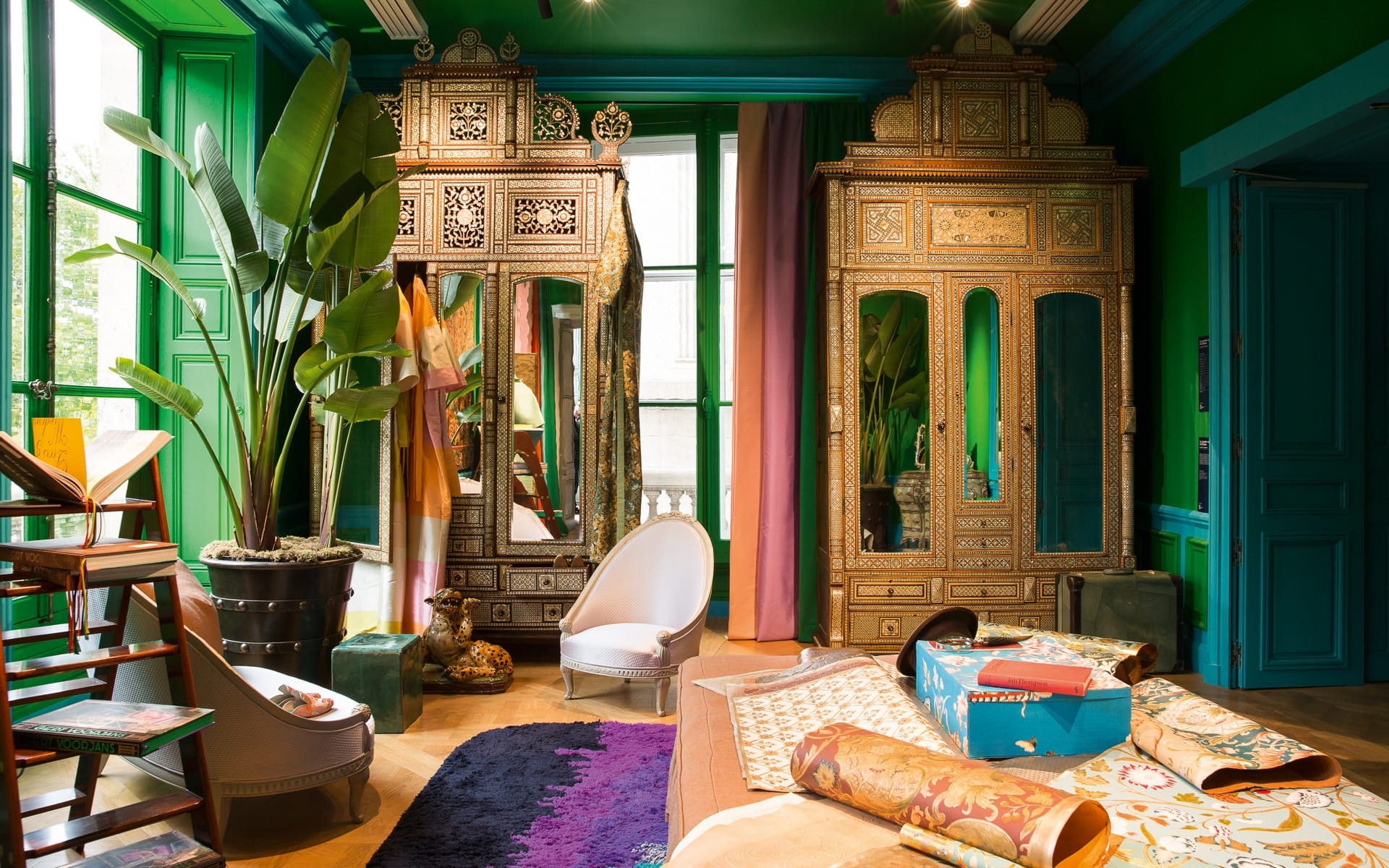 Colorful room with ornate mirrored cabinets, potted plants, and various fabric samples on the floor and chairs.