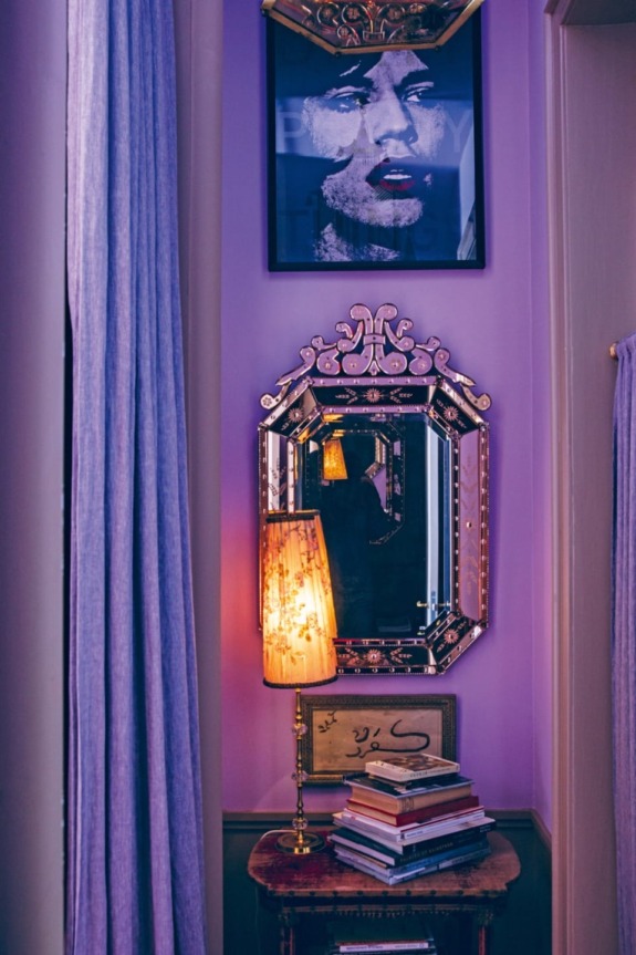 Purple room corner with decorative mirror, stack of books on a table, lamp, and artwork featuring a woman's face.