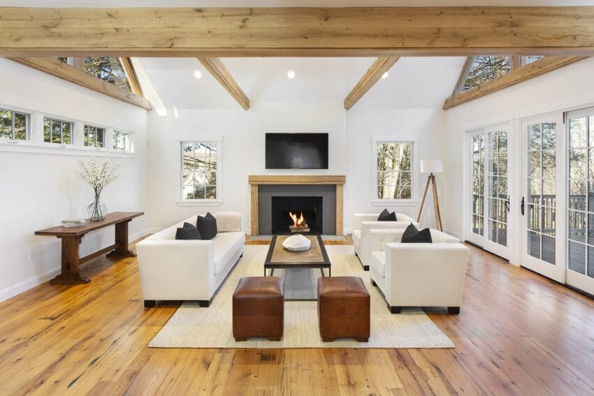 Modern living room with wooden beams, white sofas, fireplace, and large windows showcasing a forest view.