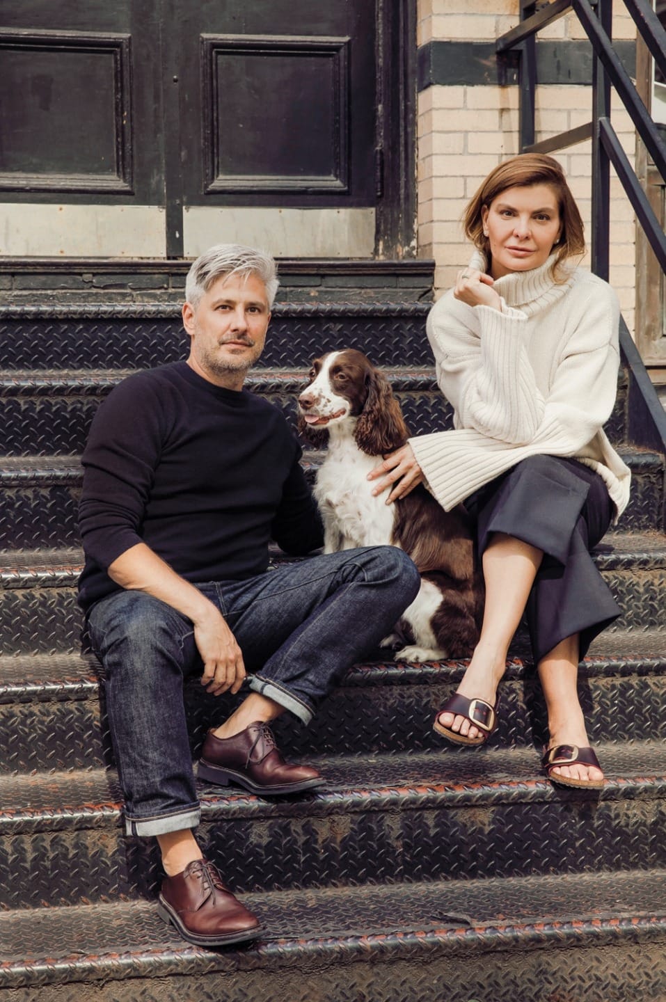 Man and woman sitting on outdoor steps with a dog, both casually dressed and looking at the camera.