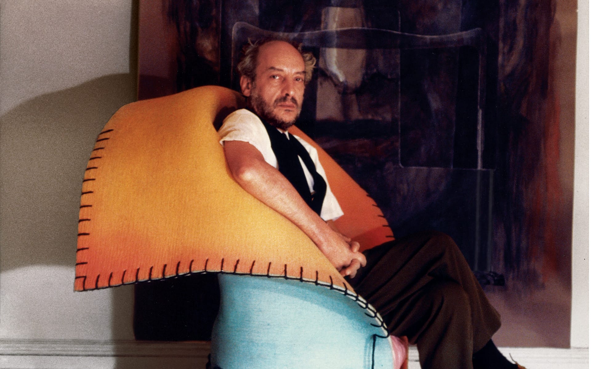 Man sitting on a colorful chair with a unique design, wearing a shirt and vest, in front of an abstract painting.
