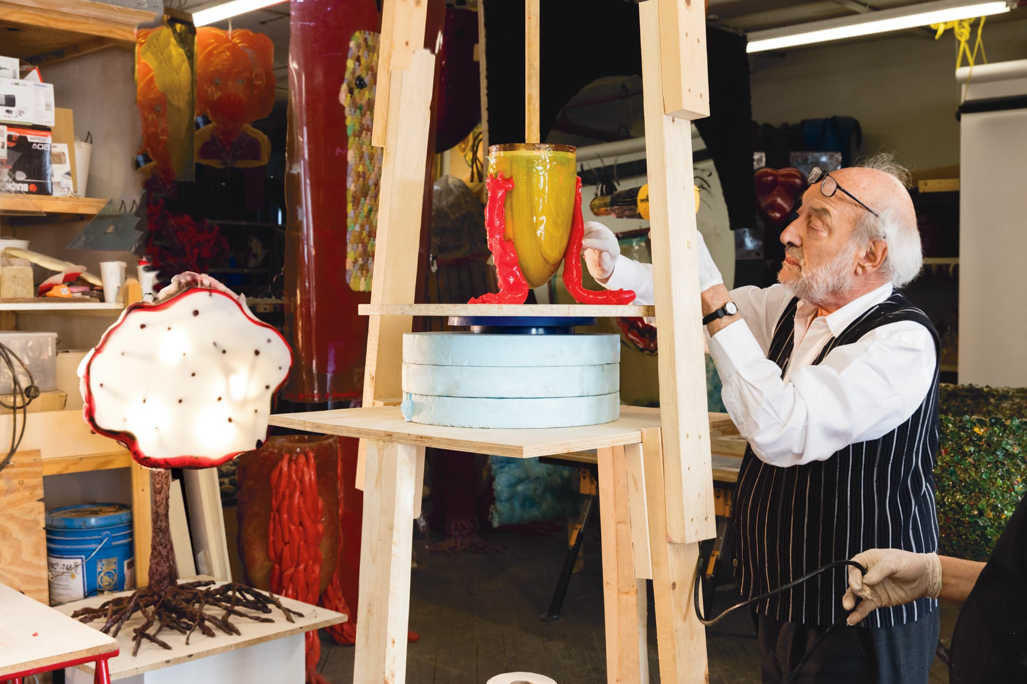 Man in workshop carefully assembling colorful sculptures on a wooden stand, surrounded by art materials and vibrant decor.