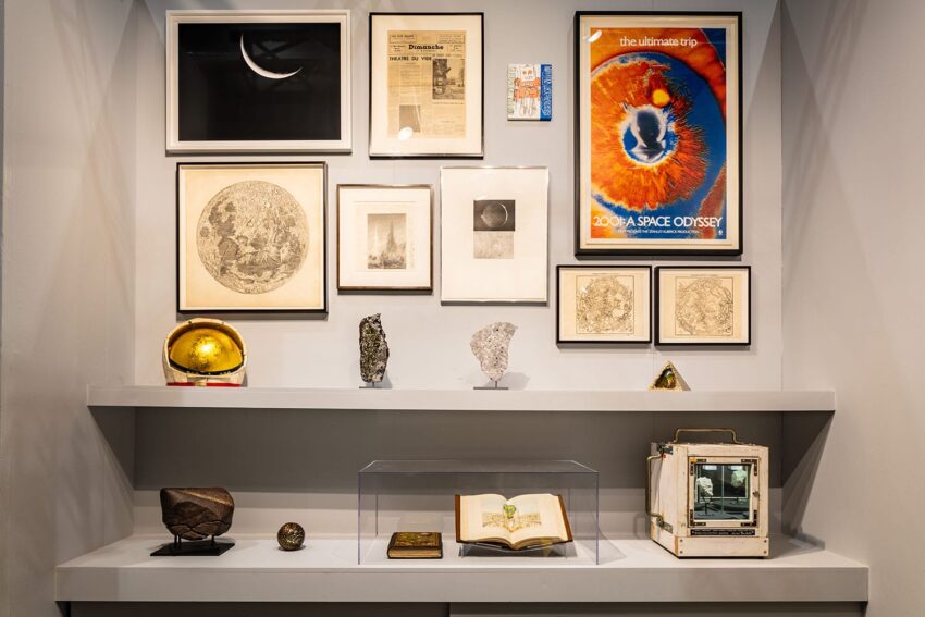 Gallery wall featuring framed art, newspaper clippings, and space memorabilia, with shelf displaying rocks and an open book.