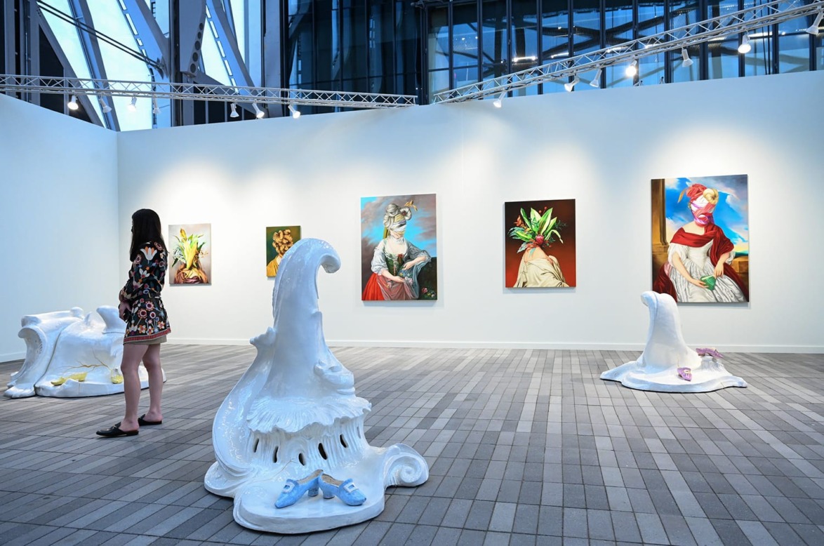 Art gallery with a woman viewing abstract sculptures and colorful paintings on a white wall under bright lighting.