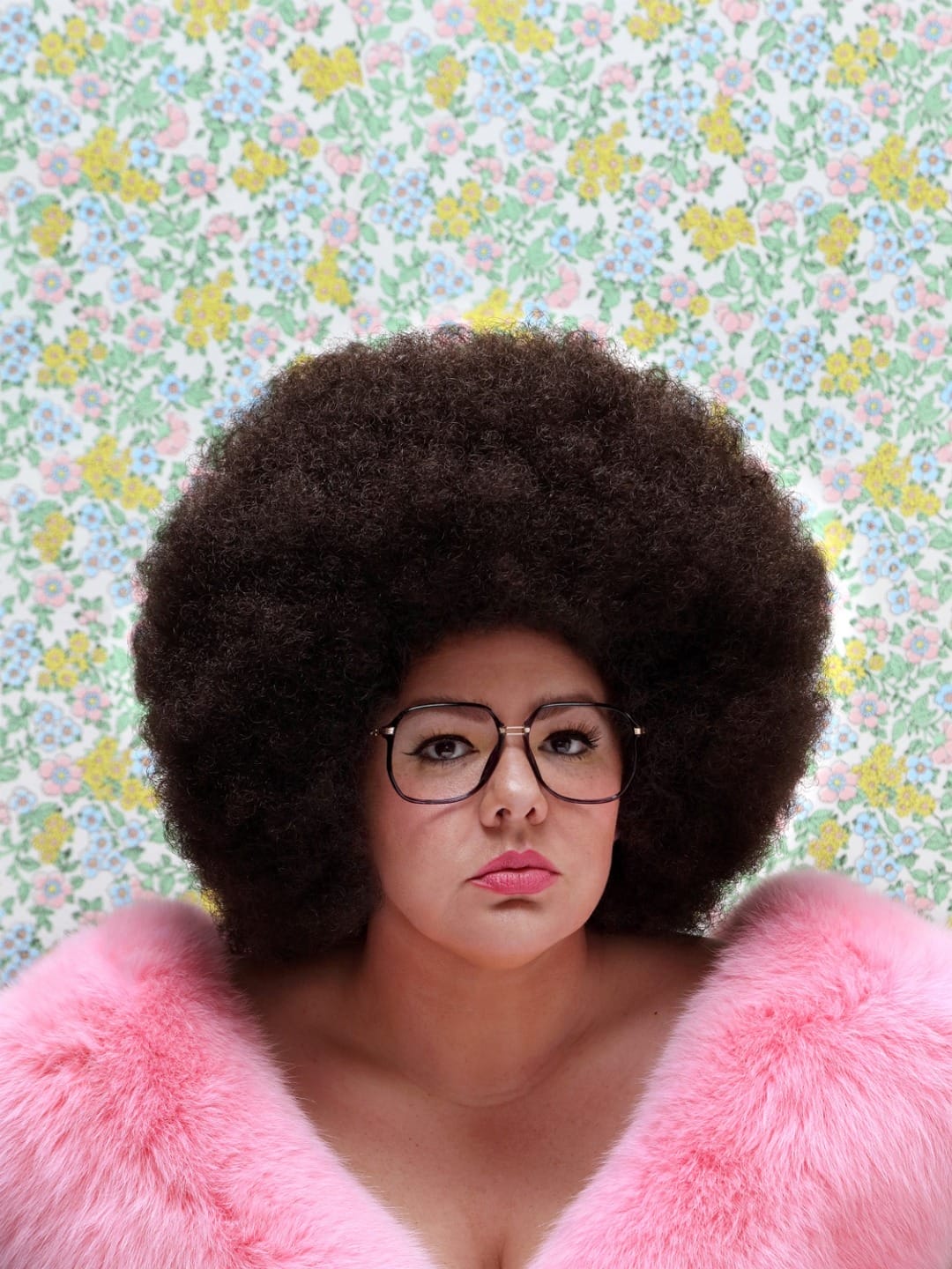 Person with large afro wearing glasses and a pink fur coat, set against a floral patterned background.