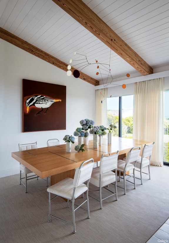 Modern dining room with a wooden table, white chairs, artwork of a bird, ceiling beams, and a colorful mobile hanging above.