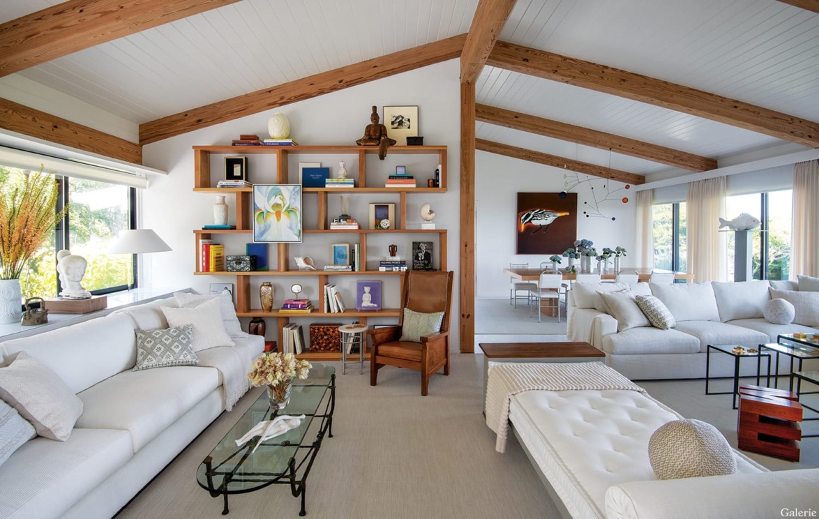 Spacious living room with white sofas, wooden beams, shelves with books and art, large windows, and modern decor.