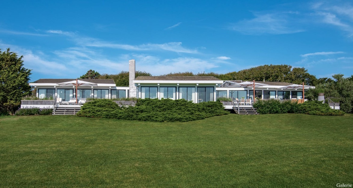 Modern hillside house with large glass windows, surrounded by greenery under a bright blue sky.