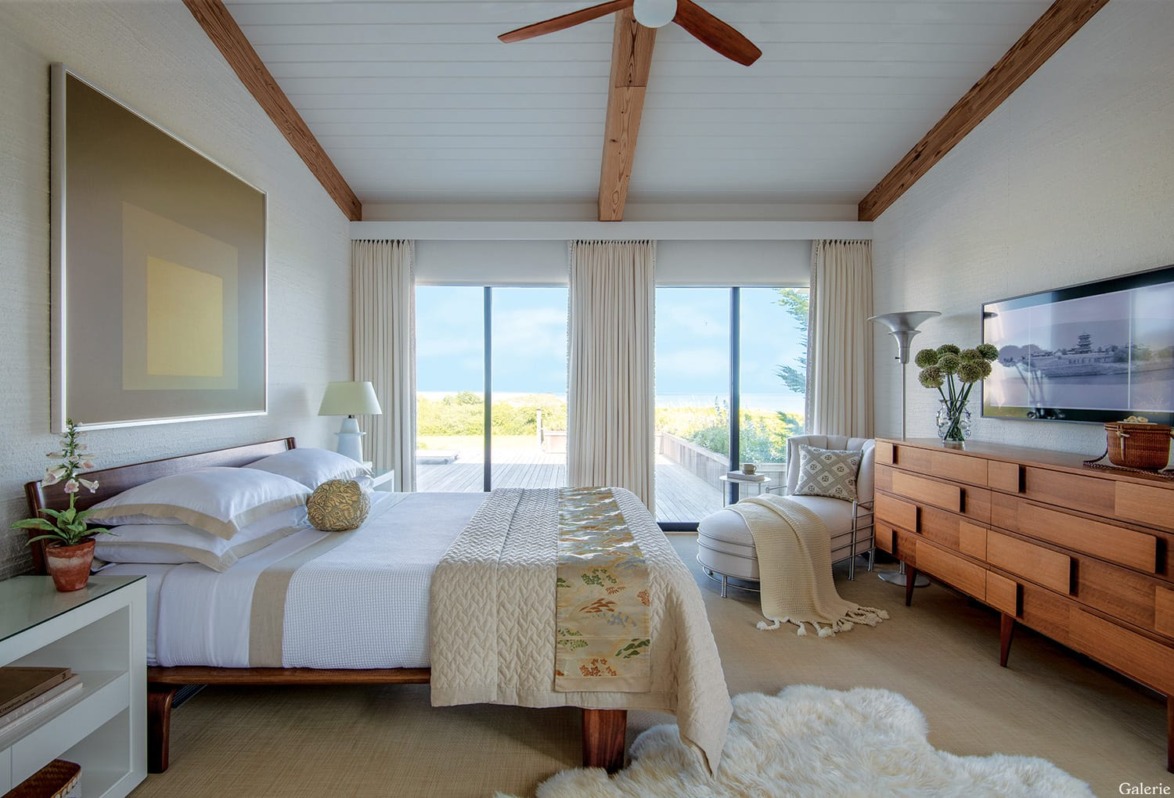 Modern bedroom with large windows, wooden ceiling beams, mid-century furniture, and a view of the ocean.