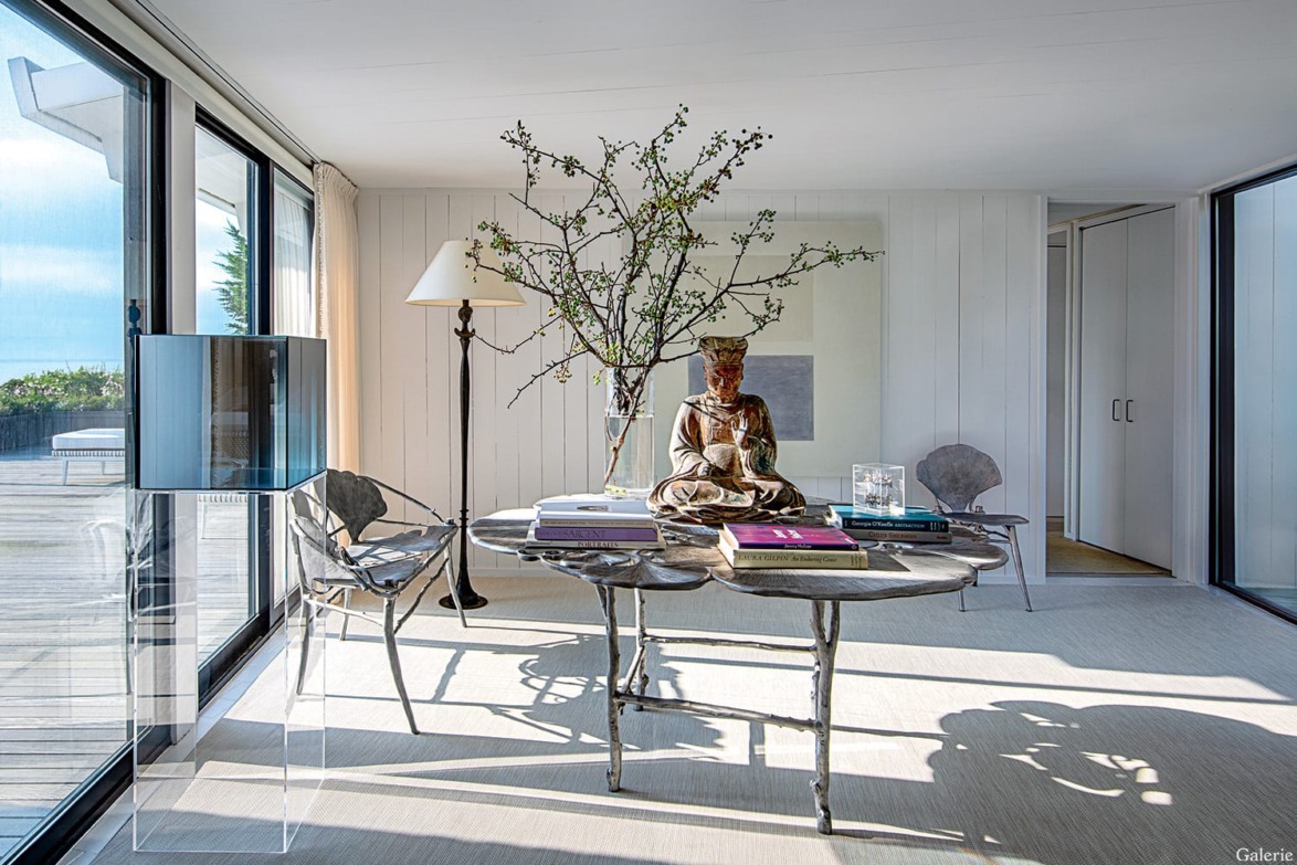 Modern living room with a metallic table, statue, and tall branches, surrounded by glass doors and natural light.