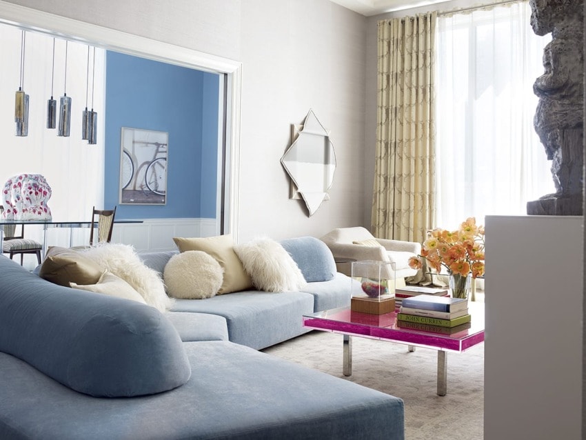 Modern living room with blue sofas, pink coffee table, decorative pillows, and wall art near a large window with curtains.