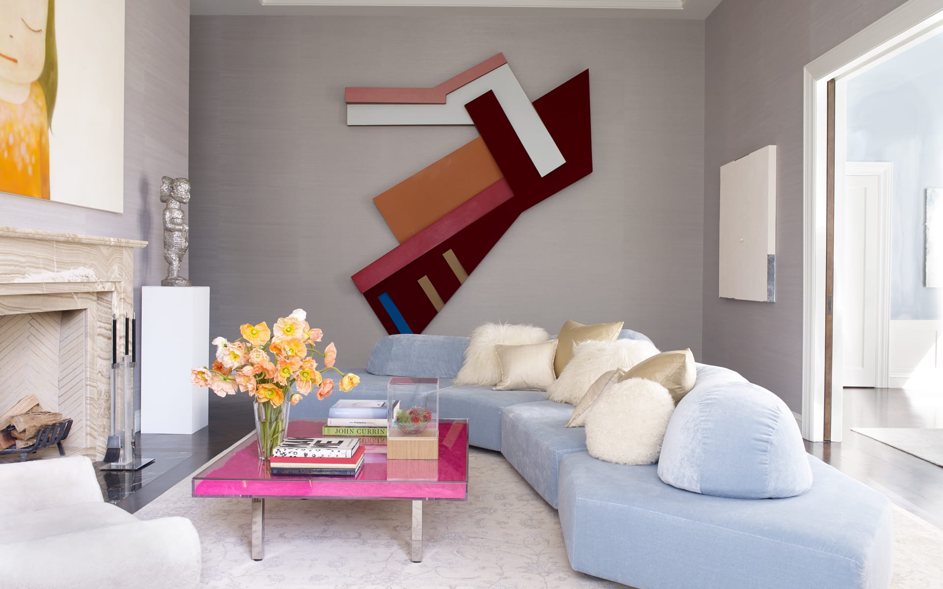 Modern living room with abstract wall art, blue sectional sofa, pink coffee table, and a vase of flowers near a fireplace.
