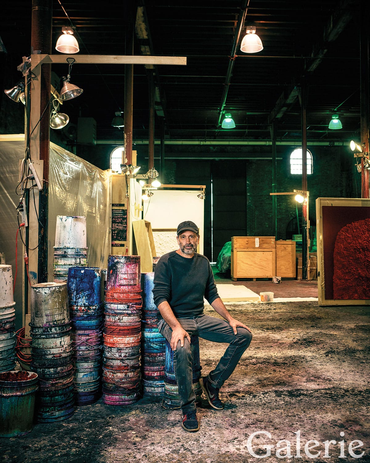 Artist seated in an industrial studio surrounded by stacked colorful paint cans.