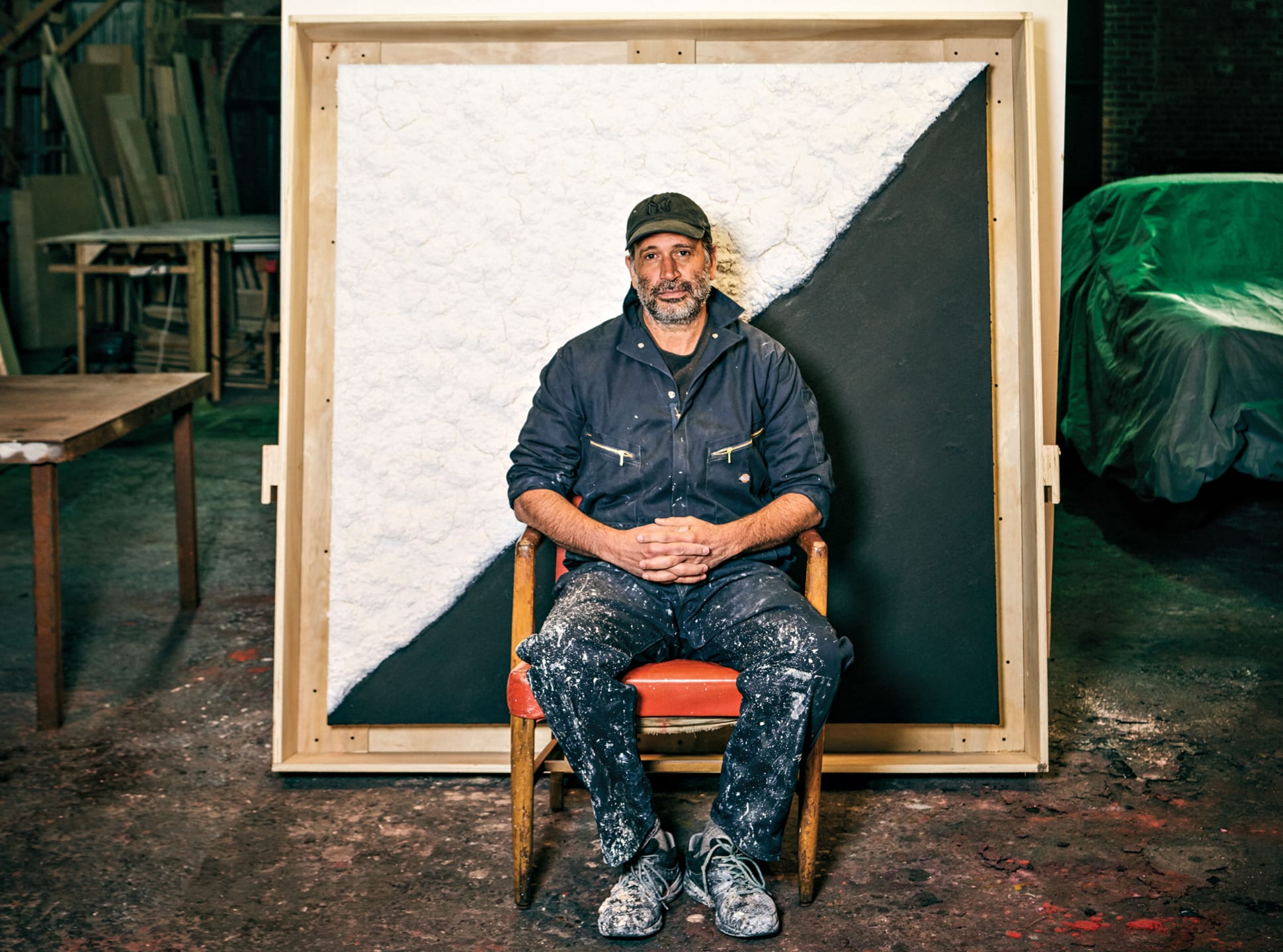 Artist seated in workshop wearing paint-splattered overalls, sitting in front of a large abstract canvas with texture.