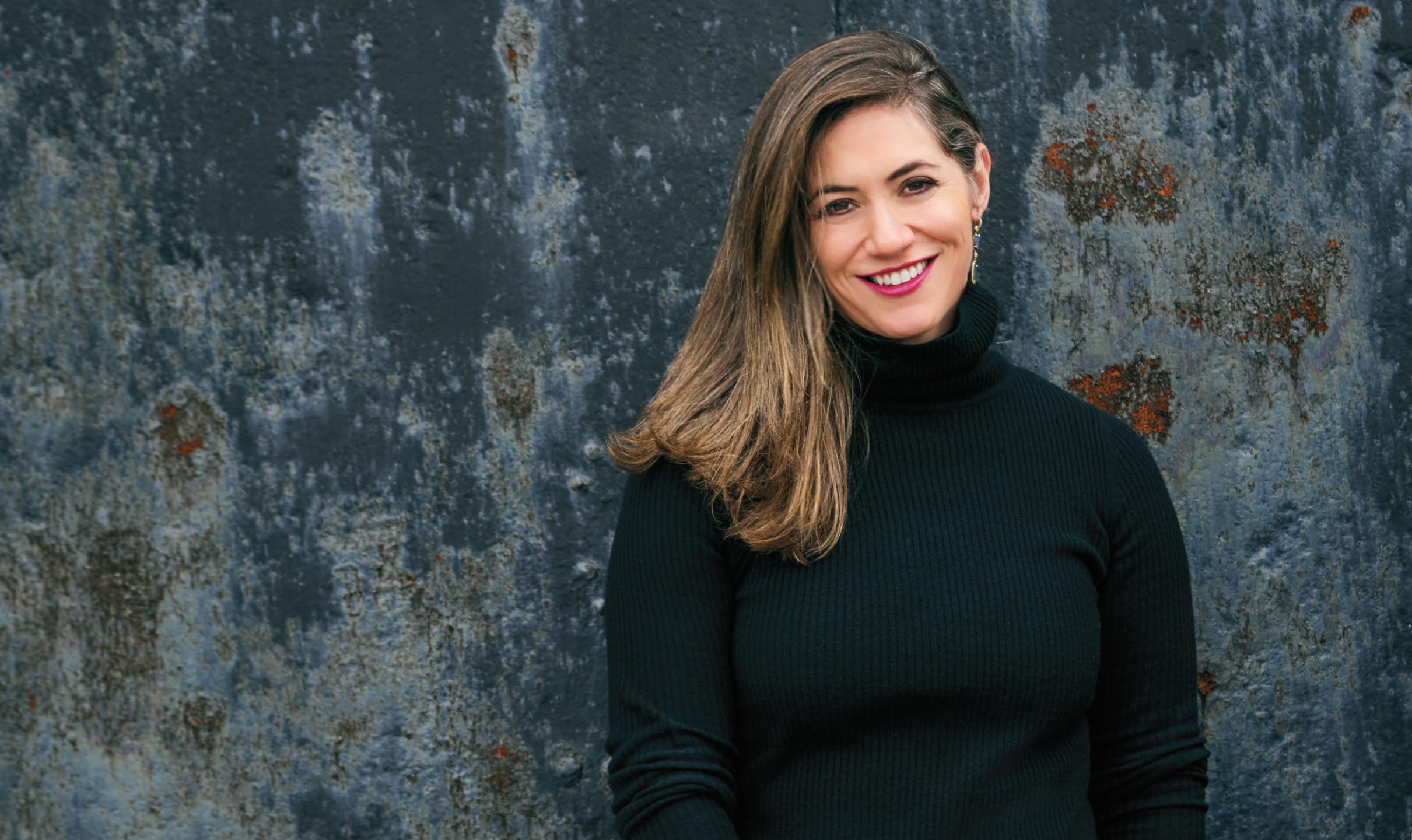 Smiling person in a black sweater standing against a textured, weathered wall.