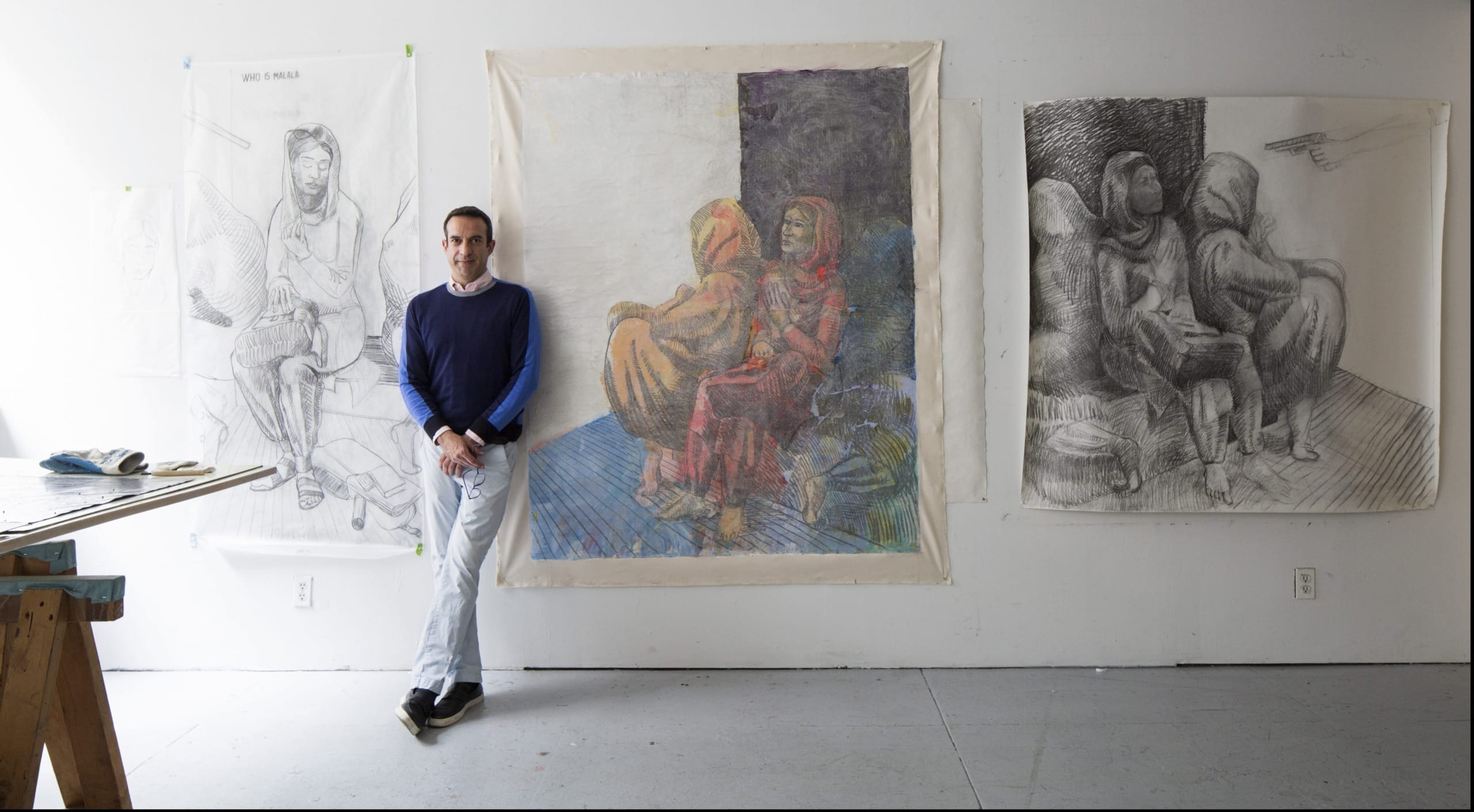 Artist standing beside large sketches and paintings in a studio with art supplies visible.