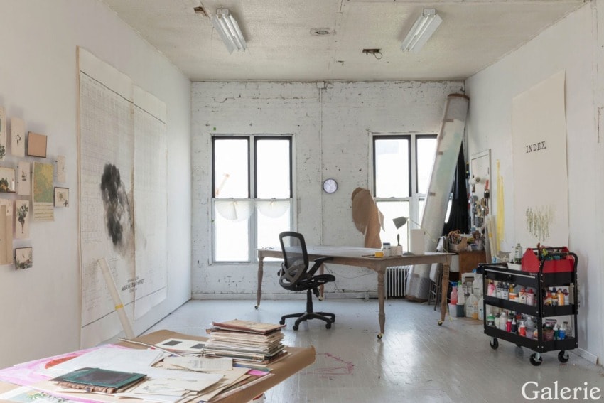 Artist's studio with a central desk, artwork on the walls, and supplies in a rolling cart by a window in a well-lit room.